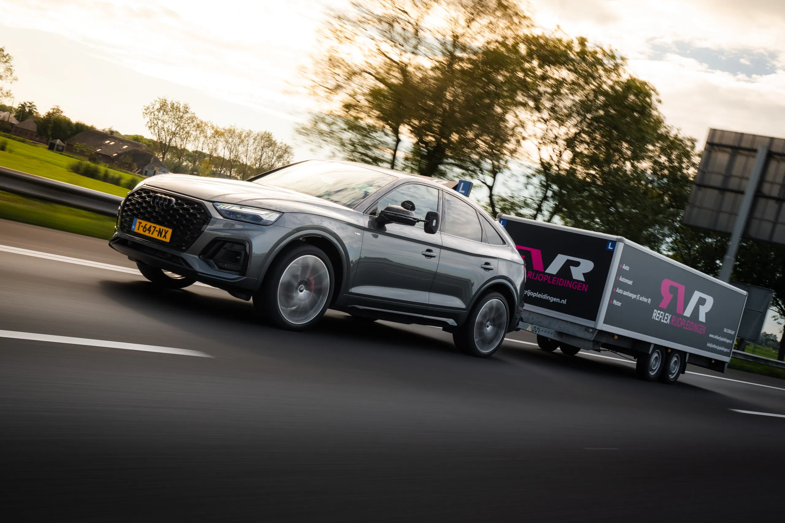 Een grijze Audi SUV lesauto van Reflex Rijopleidingen rijdt met een grote aanhangwagen op de weg tijdens een BE aanhanger rijles in regio Woerden.
